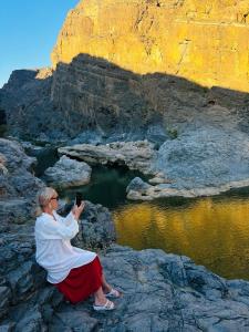 Eine Frau, die auf einem Felsen sitzt und ein Bild vom Wasser macht. in der Unterkunft Wadi Al Arbeieen Resort in Muscat