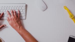 Manos escribiendo en un teclado con un reloj de pulsera y un teléfono celular al costado