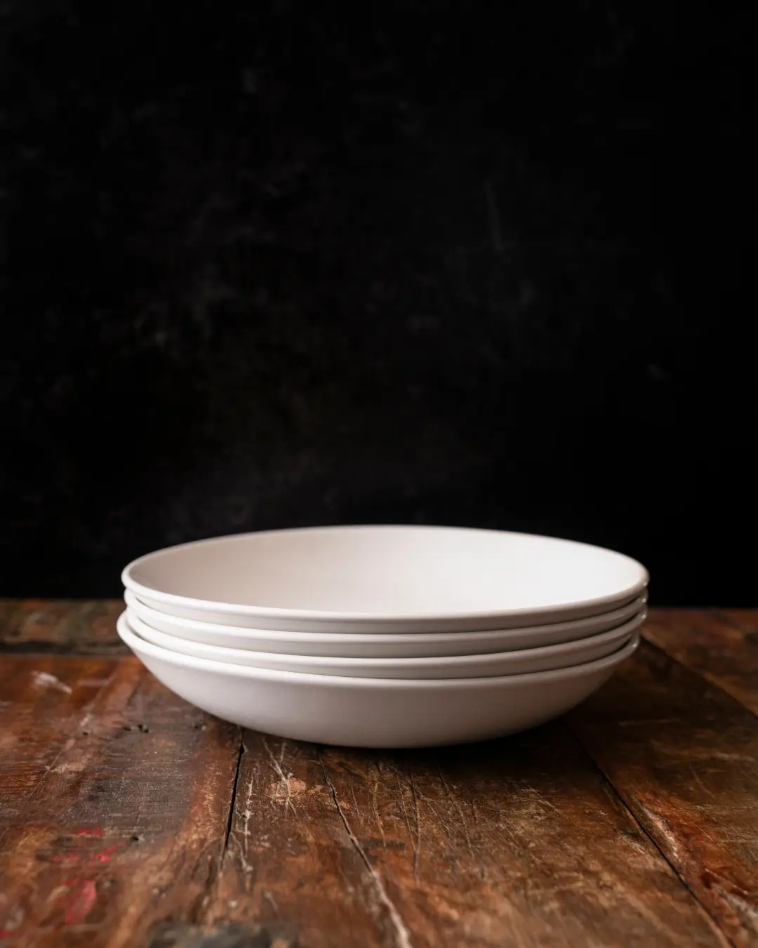 A stack of white plates is positioned on a worn wooden table against a dark background.