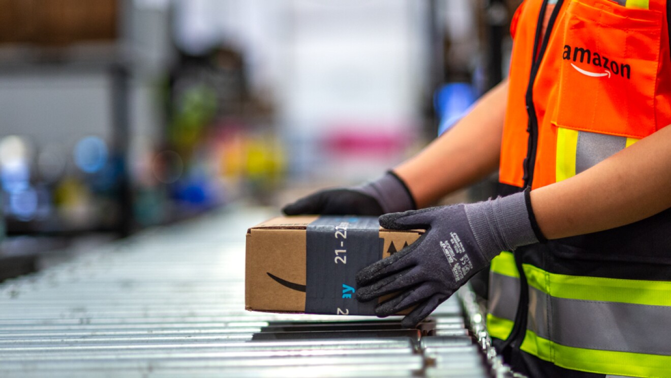 Plano corto de las manos y parte del cuerpo de un trabajador de Amazon que está agarrando una caja de la cinta transportadora metálica. La caja es marrón con la cinta de color gris. El trabajador de Amazon lleva guantes de seguridad de color gris y una chaleco de color naranja, gris y amarillo con el logo de Amazon.