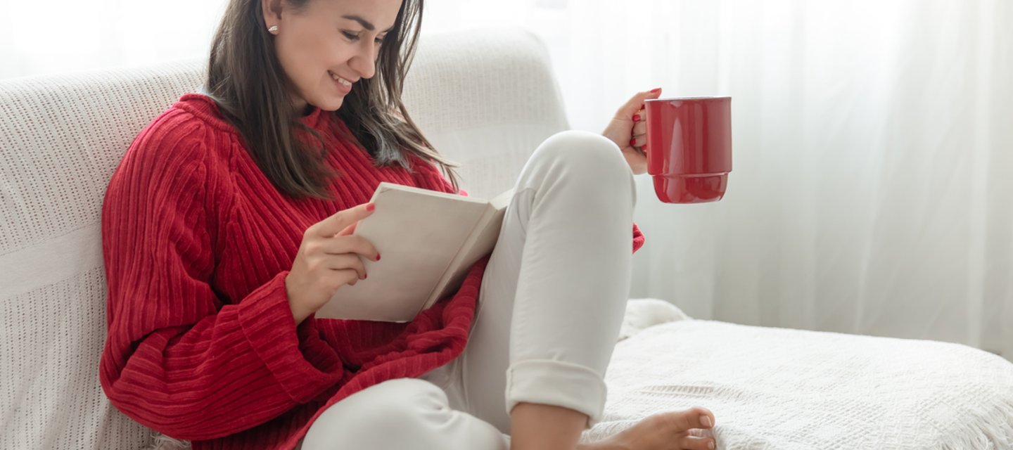 Woman in red sweatshirt and a cup in her hand reading a book on a white couch