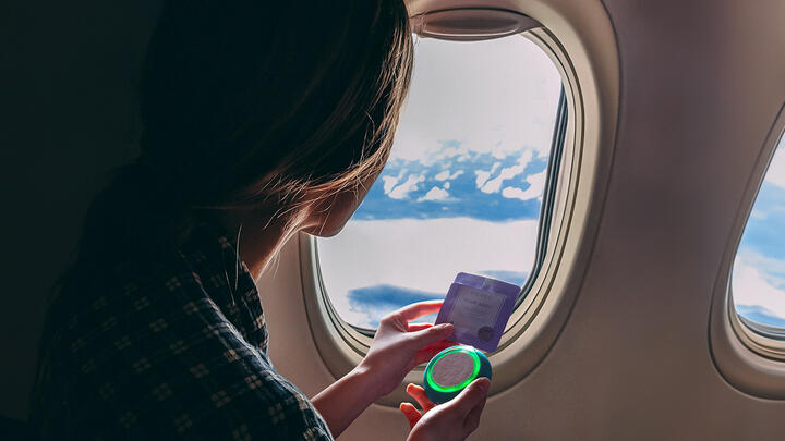 Woman sitting on a window seat in an airplane, holding FOREO UFO 3 go deep facial hydration device and accompanying FOREO mask