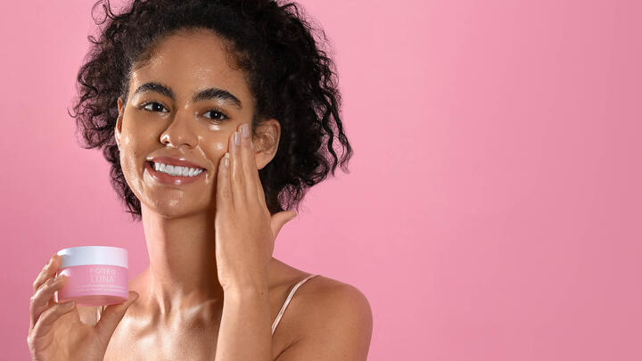 Young female model applying FOREO LUNA Ultra-Nourishing Cleansing Balm to her cheeks, standing in front of a pink background