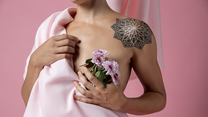 Female model wrapped in a pink sheet, her left breast exposed, holding a small bouquet of flowers in front of her breast