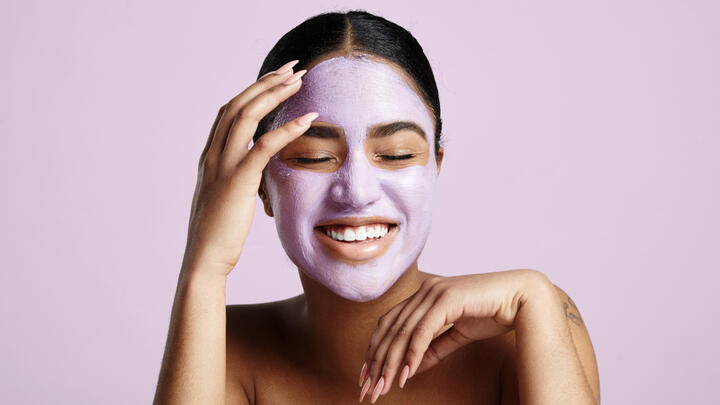 A photo of a smiling young woman with pale purple clay mask on her face in front of a lilac background