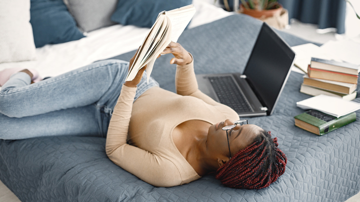 Woman reading a book in a bed, laptop and a stash of books next to her