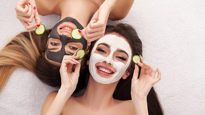 Two young woman on a bed with clay masks on their faces, holding cucumber slices