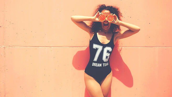 Curly woman standing in front of an orange wall in a swimsuit, holding orange halves in front of her eyes