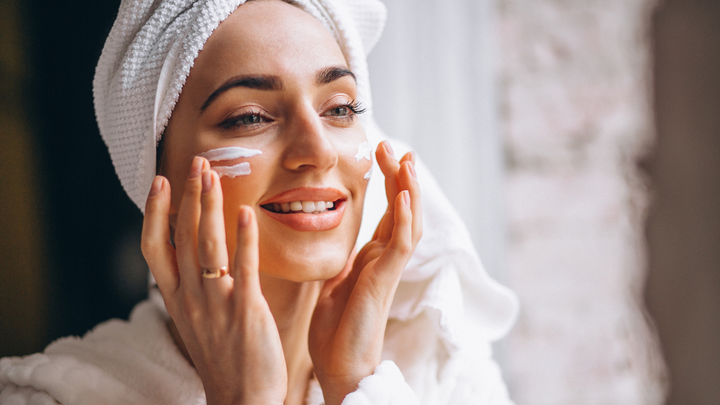 Smiling woman with hair wrapped in a towel applying moisturizer to her cheeks