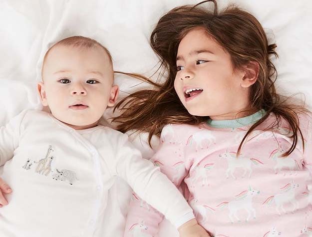 A baby in white pajamas next to a child wearing the rainbow unicorn pajama set