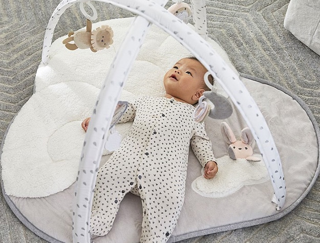 Baby in pajamas looking up at toys hanging from activity gym.