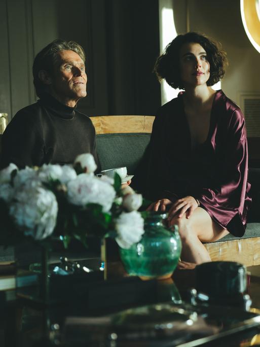 Willem Dafoe and Margaret Qualley sitzen in einer Szene des Films "Kinds Of Kindness" von Yorgos Lanthimos nebeneinander auf einem Sofa.