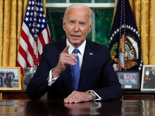 US-Präsident Joe Biden sitzt im dunklen Anzug an seinem Schreibtisch im Oval Office und gestikuliert mit seiner rechten Hand. Im Hintergrund sind Flaggen, Vorhänge und persönliche Fotos zu sehen