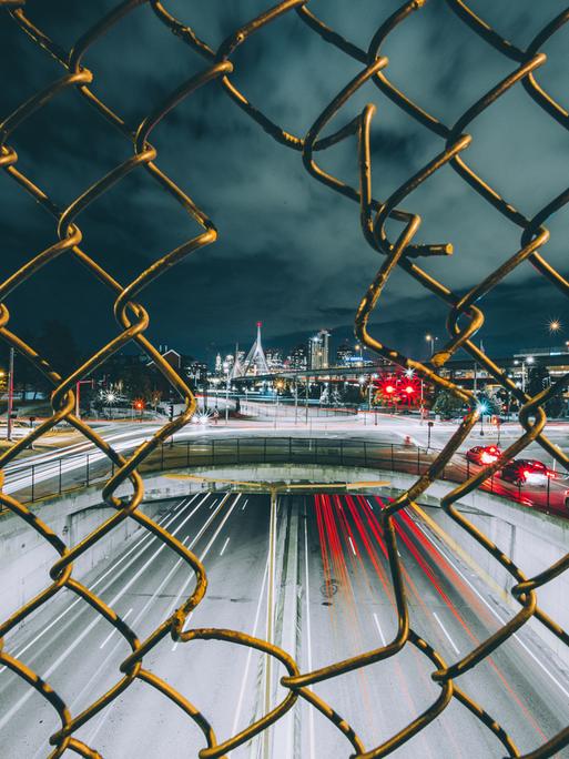 Blick durch einen Maschendrahtzaun auf eine dramatisch beleuchtete Stadt bei Nacht