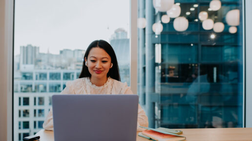 A young woman in a modern office space uses a laptop at a desk. Space for copy.