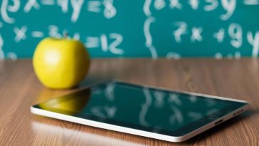 Edtech companies, yellow apple on brown table next to tablet.