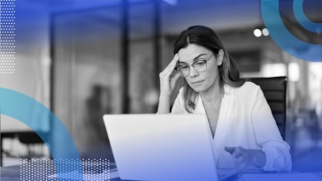 A woman wearing glasses and a weary expression is looking at a computer screen.