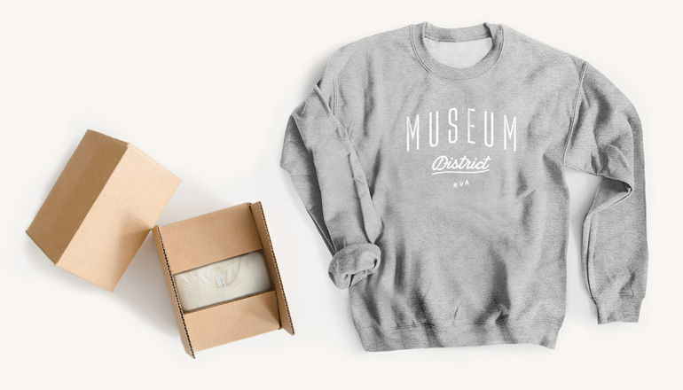 An unfolded, gray crewneck sweatshirt with a custom design on laying next to a couple of opened cardboard boxes containing more shirts.