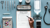 a doctor holds a pen over a clipboard. Various medical supplies are organized neatly on the table around the clipboard including a thermometer, an IV bag and a stethoscope.