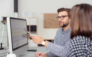 Two business professionals sit at a desk and look at a desktop computer monitor while one of them gestures to the screen with a pen.