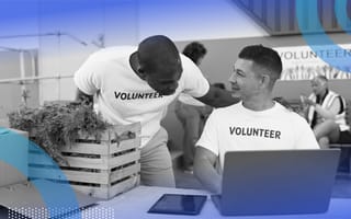 A person wearing a volunteer t-shirt is sitting at a computer and talking to another volunteer.