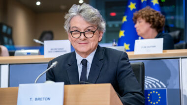 Ein Mann im Anzug mit grauen Haaren und runder Brille lächelt in die Kamera, im Hintergrund eine EU-Flagge