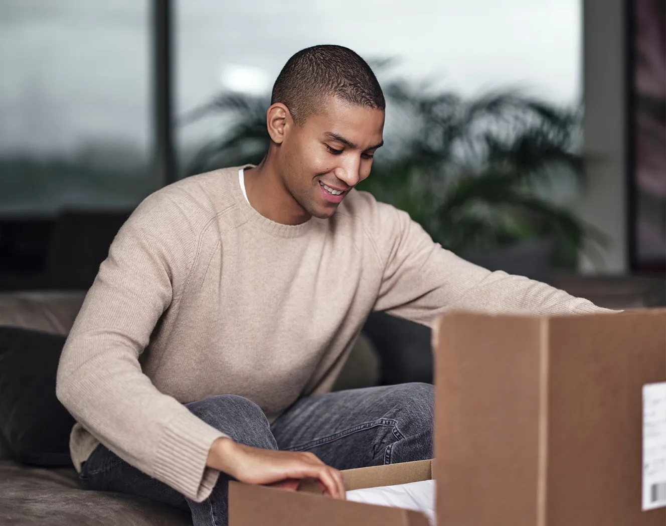 Young man sitting on a sofa, smiling, looking down and opening a cardboard box. Photo.