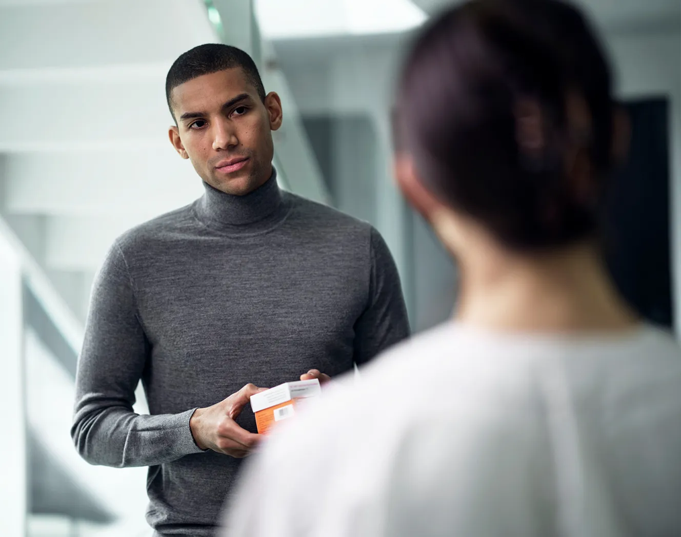 A young man, wearing a gray turtelneck pullover, with a serious look, holding a small box in his hands. He is facing a woman. The woman has her back towards us. Photo. 