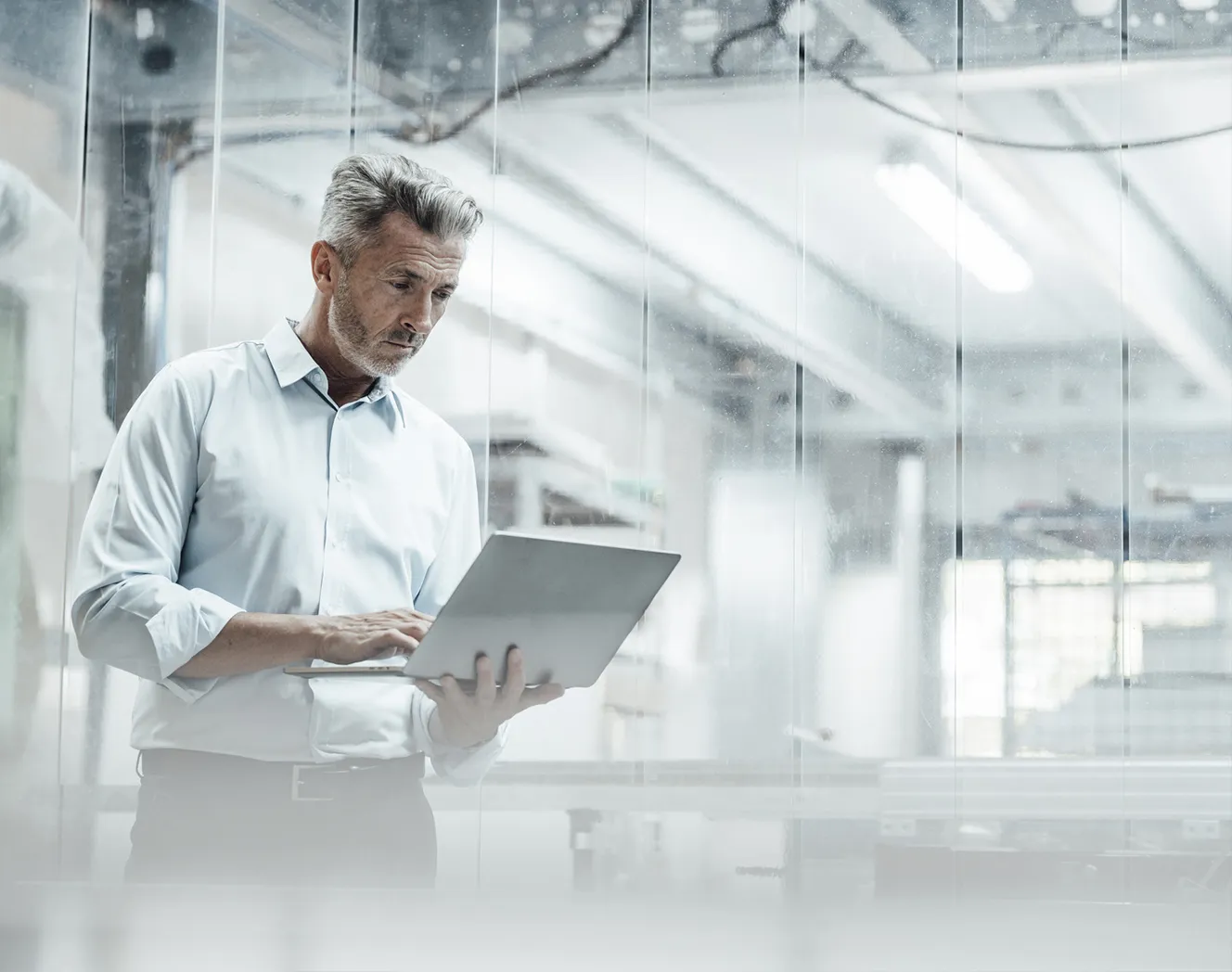 A middle aged man, wearing a white shirt, standing in a production setting. He is holding his laptop in his left hand and navigating on his laptop with his right hand. Photo. 