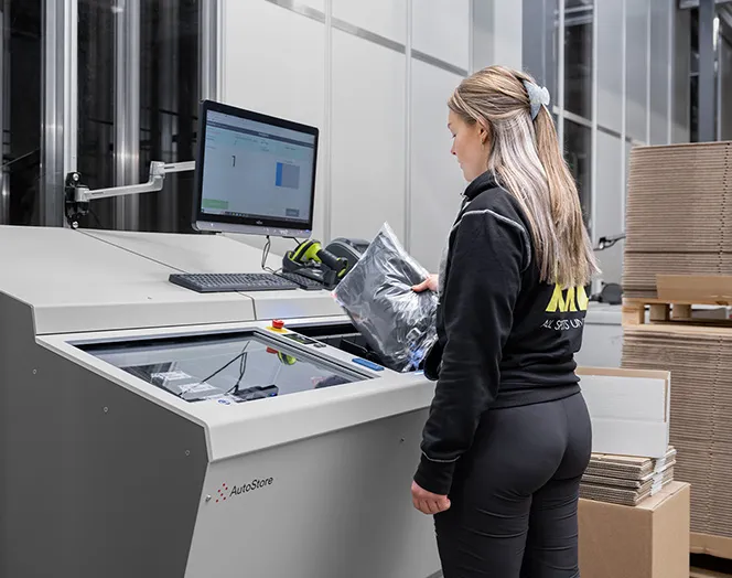 Woman standing in front of an AutoStore FusionPort. Location is at a warehouse. The woman is wearing a black sweatshirt with a logo on the back saying XXL Sports.