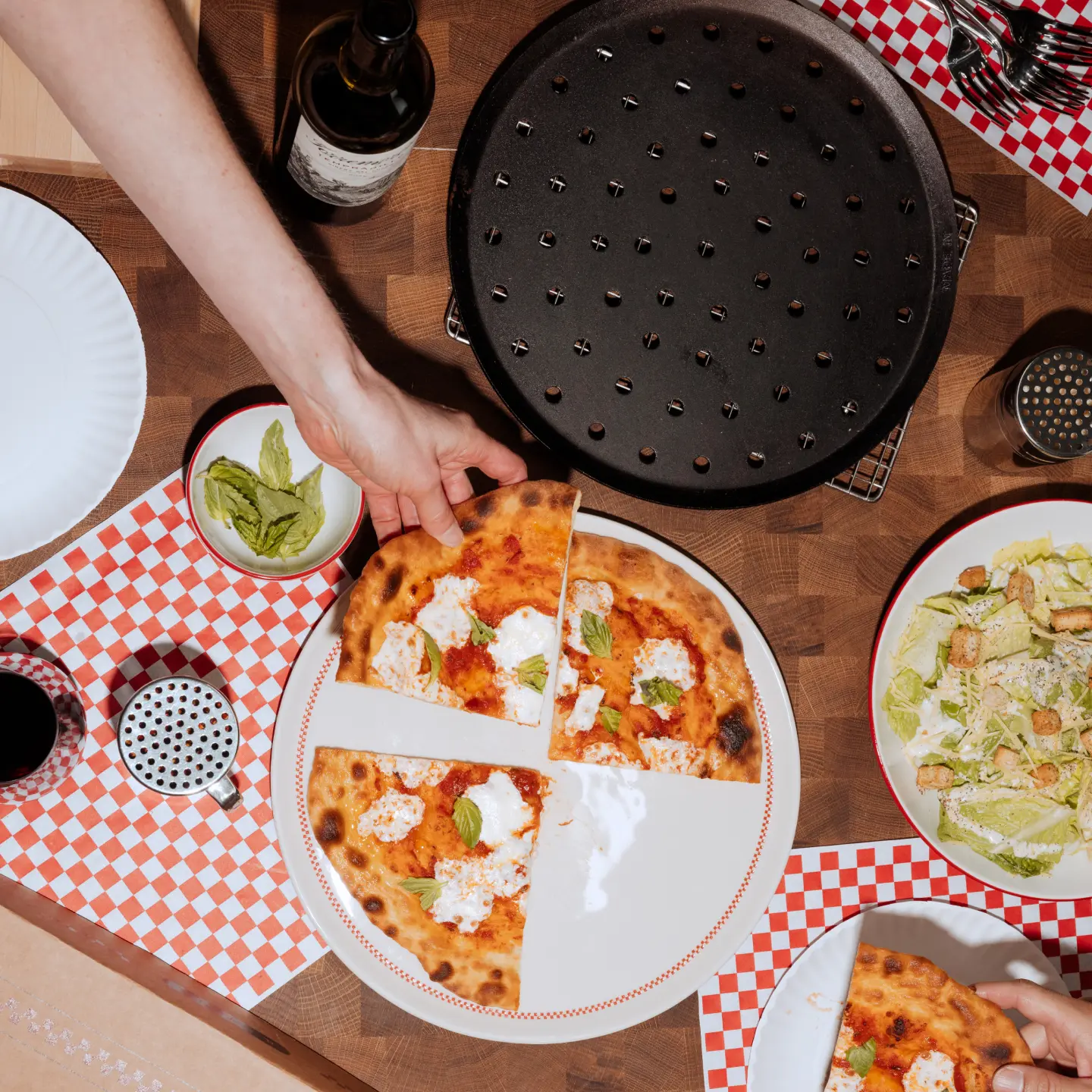 A person's hand is serving a slice of pizza from a full pie on a checkered tablecloth with salad and drinks nearby.