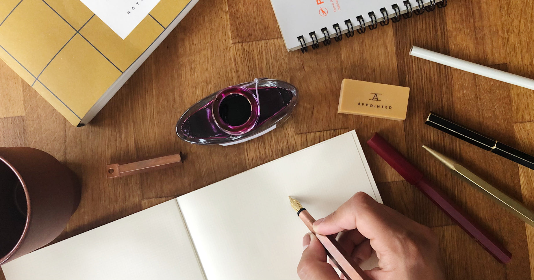 A hand gripping a fountain pen hovers above a blank notebook on a table. An inkwell, other pens, and some notebooks also rest on the table.