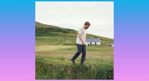 A blonde man walks through tall grass in Chelsea boots, a white tee, and blue slacks.