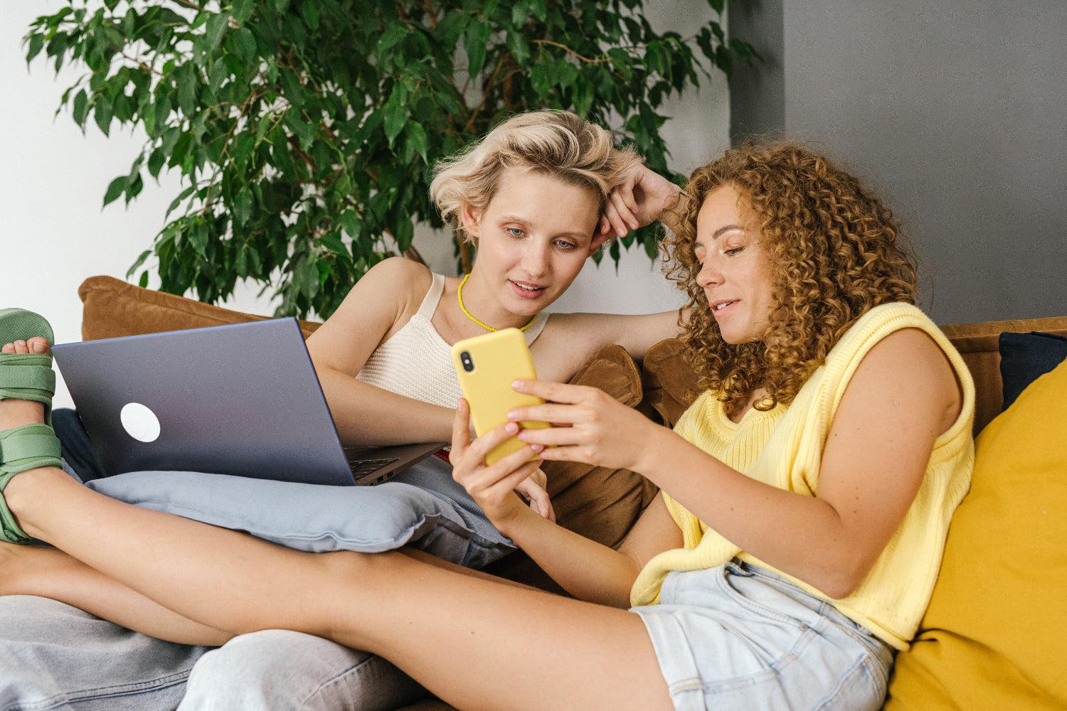 Two women look at a mobile phone together while sitting on a couch