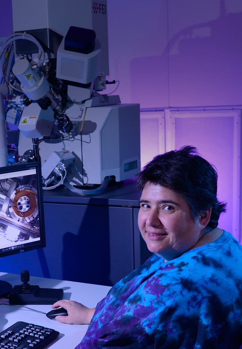 A women looks at the camera and smiles as she work at two monitors in a blue-lit room.