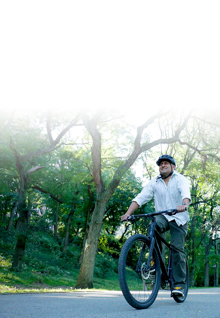 A man rides a bike wearing a helmet. Green trees are in the background.