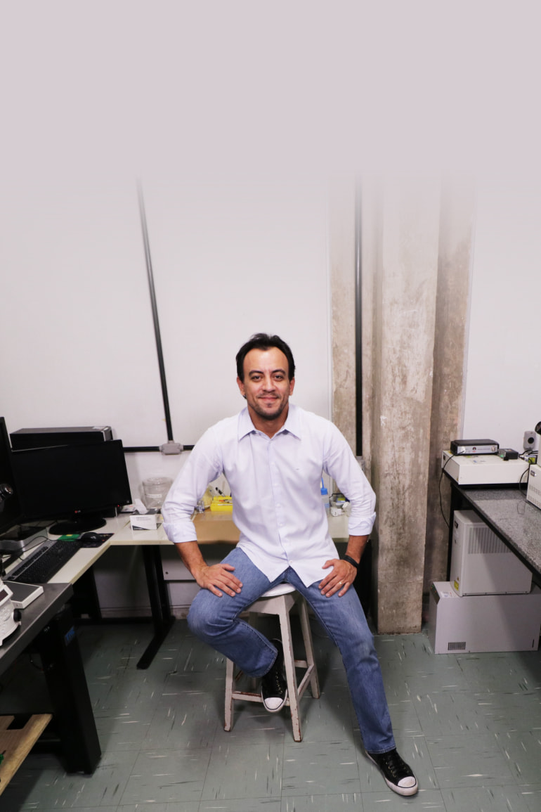 Biologist Gustavo B. Menezes of Brazil sits on a stool in a lab, with microscopes and computer screens.