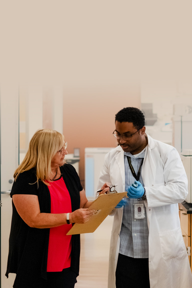 Dr. Monica Ferrini stands next to a researcher in a lab coat. Both are looking at a clipboard and in discussion with each other. They are in a research lab.