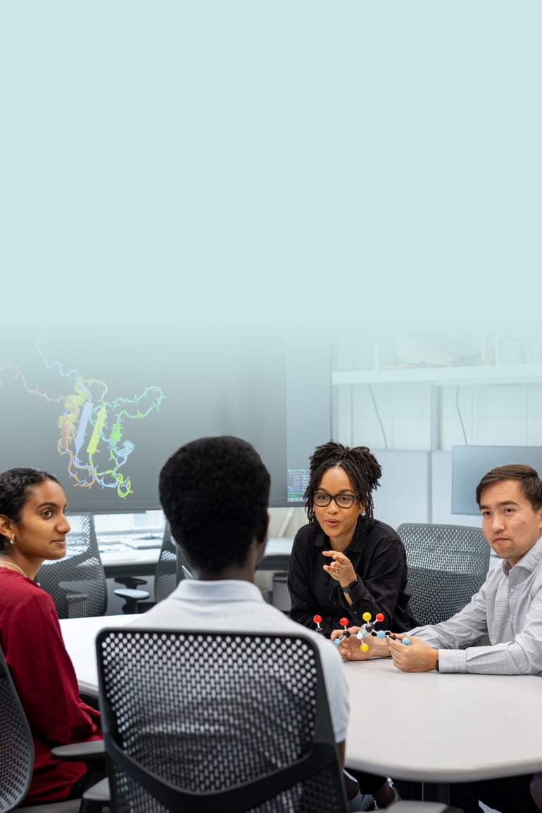 Group of researchers is sitting together at a table in a lab and talking with each other. In the background, a screen is shown depicting a photo of a protein.