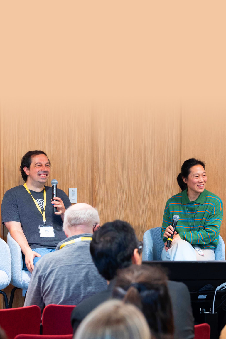 Two people are seated on a panel, both holding microphones and smiling; the man on the left, Boris Power, is wearing a T-shirt with a logo and a lanyard, while the woman on the right, Priscilla Chan, is wearing a green striped shirt. An audience is visible in the foreground.