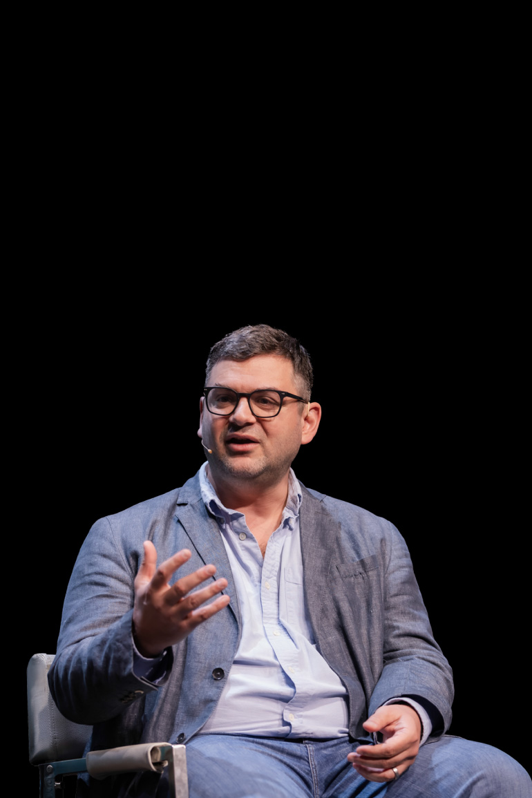 Theofanis is seated on stage and gestures to the audience with his hand. He wears a suit jacket, jeans, and glasses.