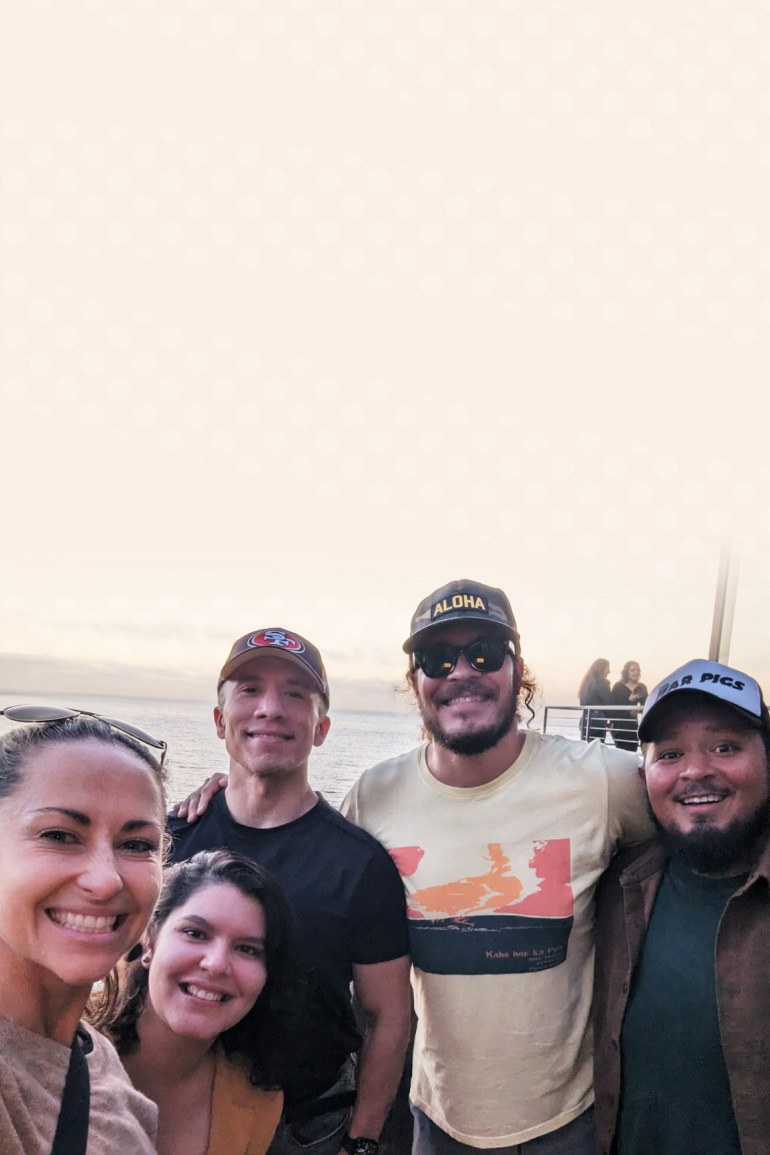 Five people smile at the camera with an ocean sunset in the background.