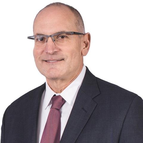 A photo of Ron Klemencic, wearing a black suit, a red tie, and glasses.