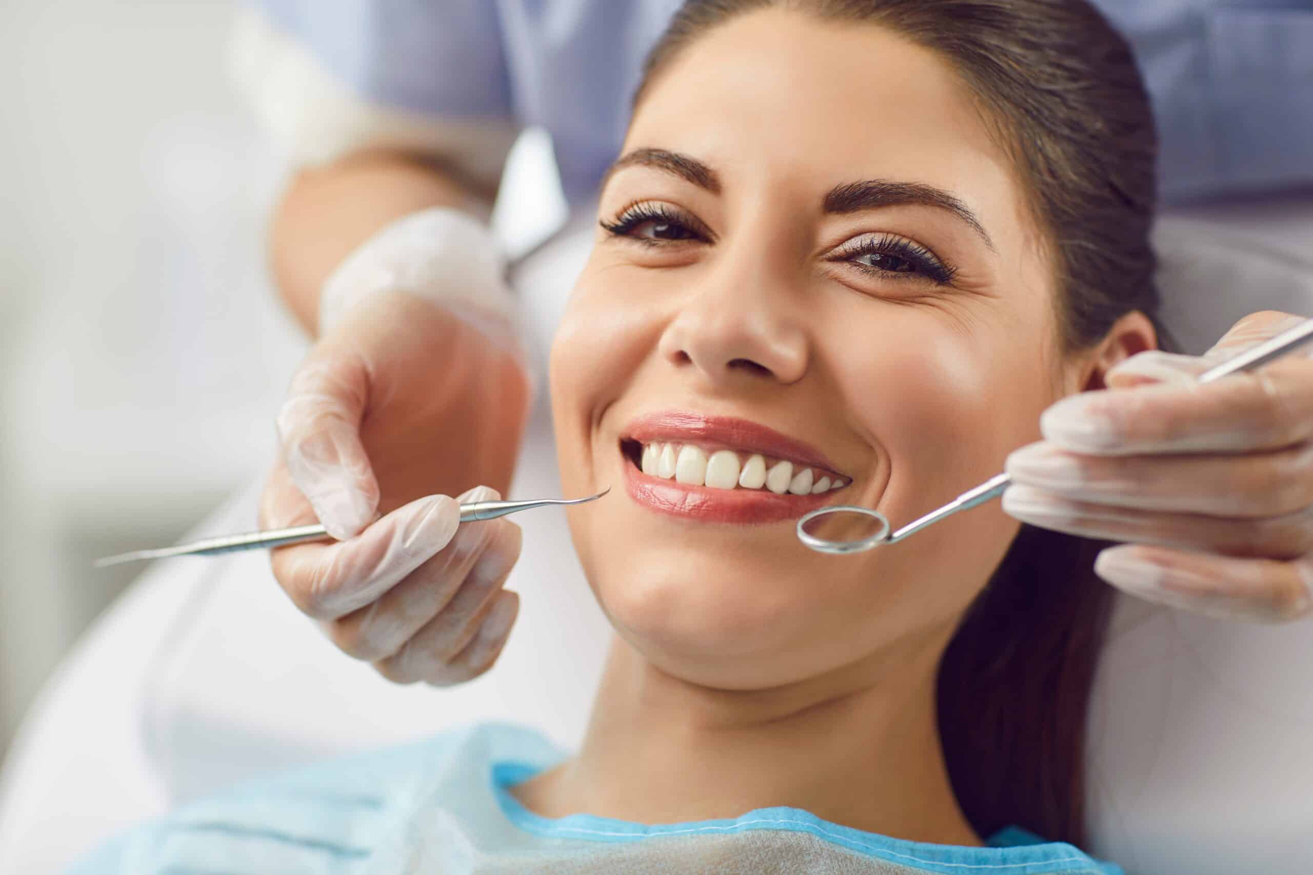 Brunette woman smiling while sitting in dental chair with dentist's hands holding teeth cleaning tools.