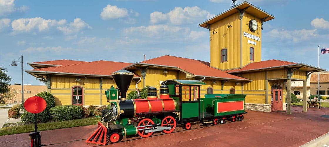 Exterior view of the yellow Dental Depot dental office in Highland Village with train in foreground.