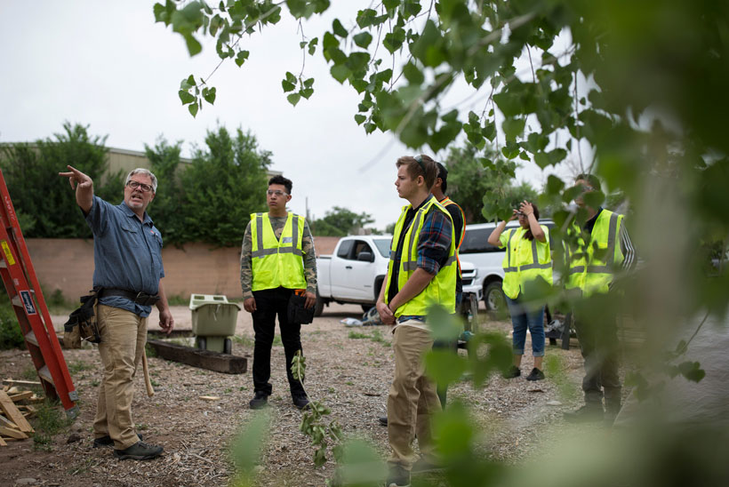 A group of 5 students wearing vests standing outside listening to instructor