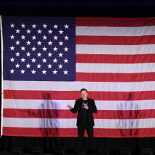 Elon Musk stands on stage in front of a giant American flag.