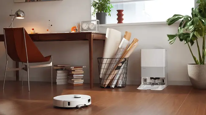 White Roborock Q Revo robot vacuum cleaning hardwood floor with dock, desk, and potted plant in background