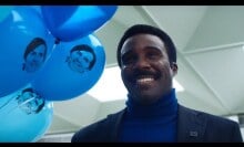 Milchick from "Severance" smiling while holding blue balloons with Mark S.'s face on them.