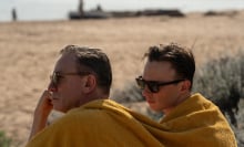 Daniel Craig and Drew Starkey sit huddled under one yellow towel on the beach in a still from the film "Queer."